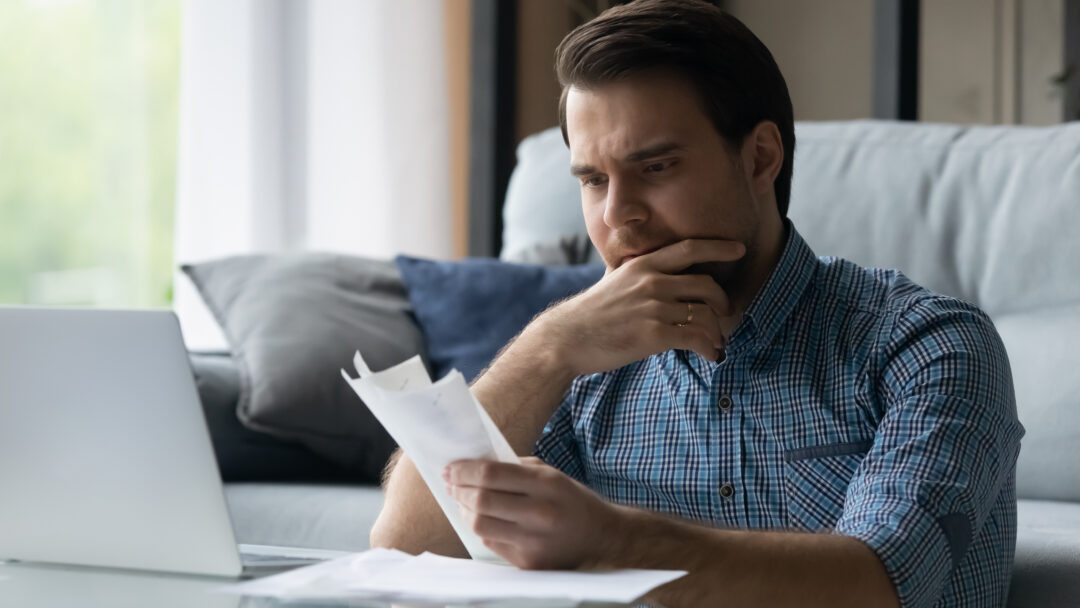 Man sitting at home reviewing bills with a worried expression while using a laptop.