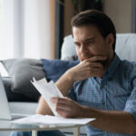 Man sitting at home reviewing bills with a worried expression while using a laptop.