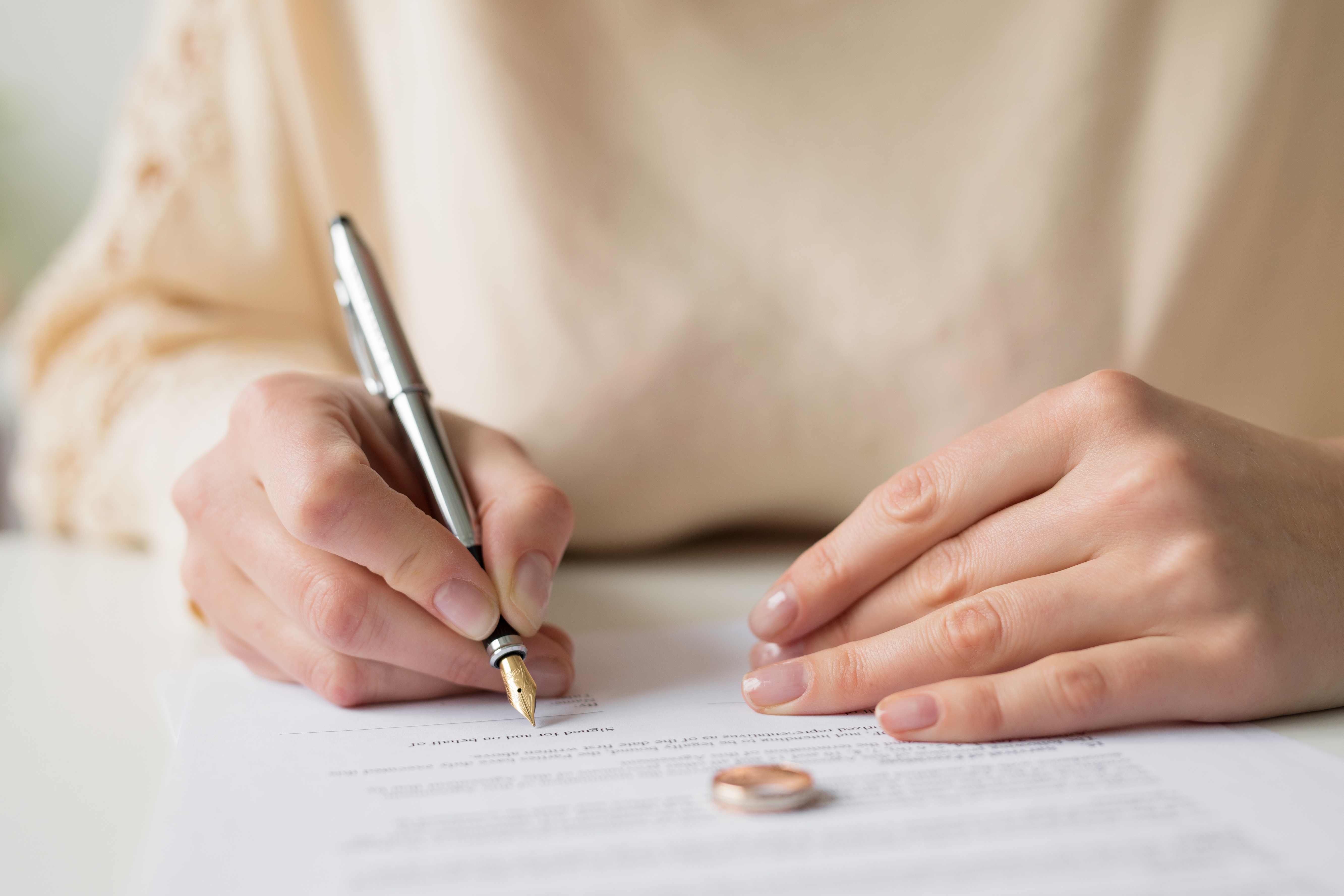 Hands holding a pen and signing legal paperwork, with a wedding ring resting on the paper.