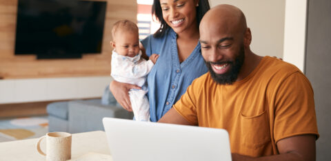 Smiling parents with their baby looking at a laptop together in the kitchen.