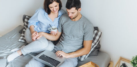 Wife and husband make shopping online, choose present for their little child in internet, look at laptop computer, enjoy togetherness in bedroom, drink hot beverage. People, leisure, technology