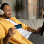 Cheerful man with laptop and credit card lying on couch, wearing headphones, shopping online at home