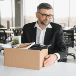 A man with glasses looking down sadly at a box of office items, suggesting he has been laid off or is leaving his job.
