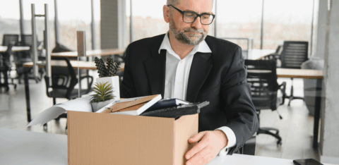 A man with glasses looking down sadly at a box of office items, suggesting he has been laid off or is leaving his job.