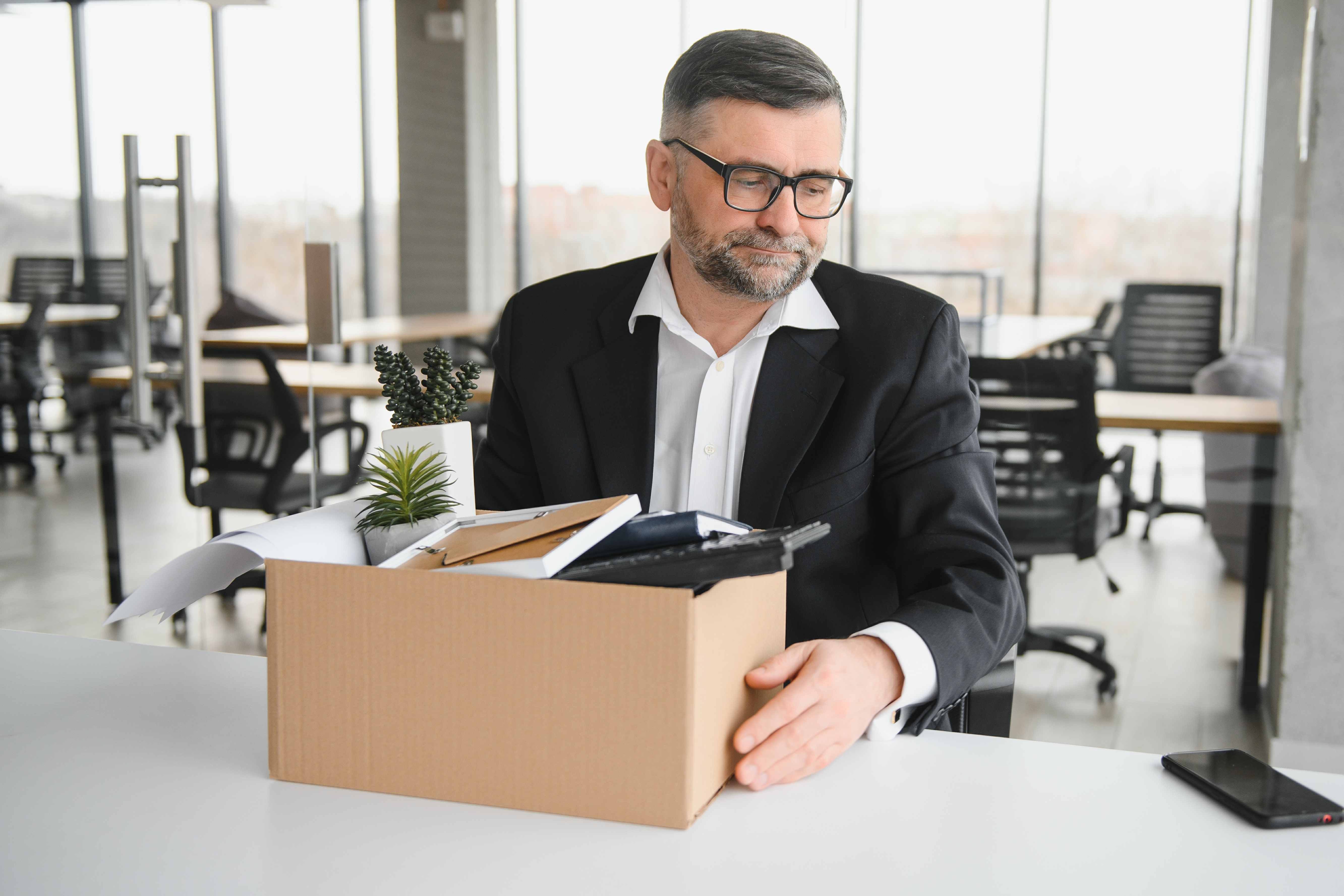 A man with glasses looking down sadly at a box of office items, suggesting he has been laid off or is leaving his job.