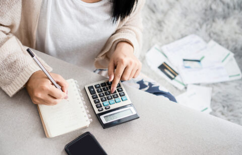 Person using a calculator and writing in a notebook, with bills and documents scattered on the floor in the background.