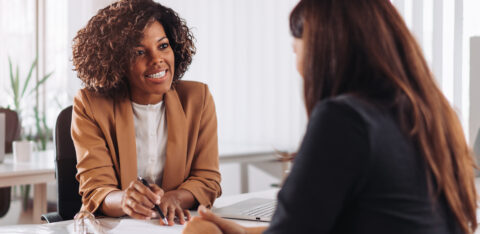 A professional woman in a tan blazer smiling while speaking with a client across a desk in a modern office setting.