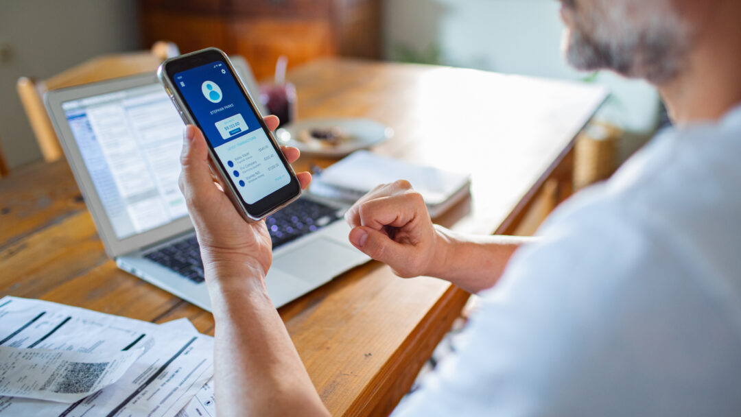 Person reviewing digital banking app on mobile phone with bills, receipts, and an open laptop on a wooden desk.