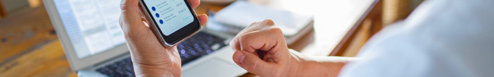 Person reviewing digital banking app on mobile phone with bills, receipts, and an open laptop on a wooden desk.