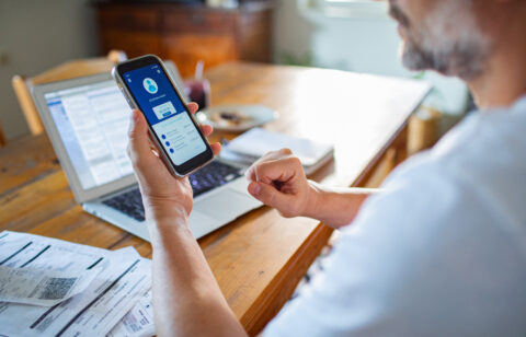 Persona revisando una aplicación de banca digital en el teléfono móvil con facturas, recibos y un ordenador portátil abierto sobre un escritorio de madera.