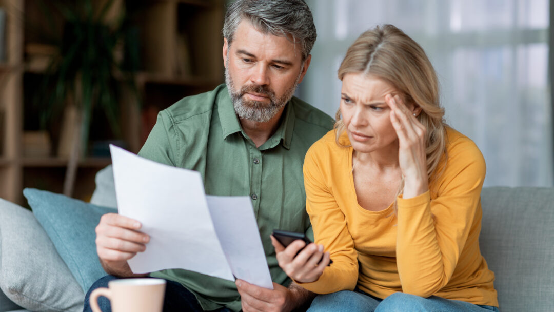 Two adults examine a document together at home, appearing worried as they check information on a phone.