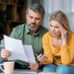 Two adults examine a document together at home, appearing worried as they check information on a phone.