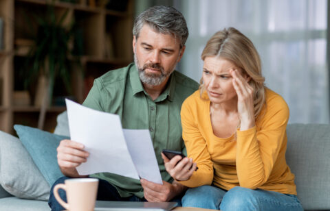 Two adults examine a document together at home, appearing worried as they check information on a phone.