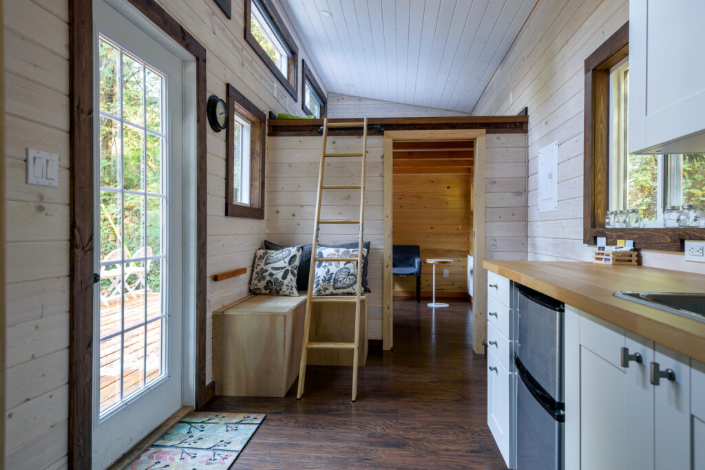 Interior of a small, modern tiny house with a loft ladder, wood-paneled walls, and a compact kitchen.