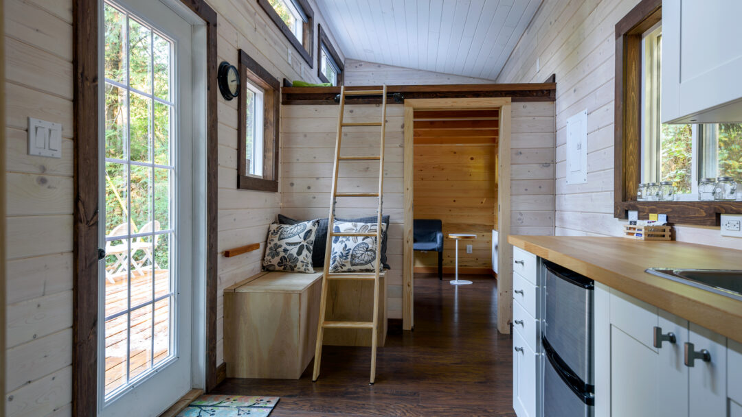 Interior of a small, modern tiny house with a loft ladder, wood-paneled walls, and a compact kitchen.