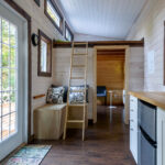 Interior of a small, modern tiny house with a loft ladder, wood-paneled walls, and a compact kitchen.