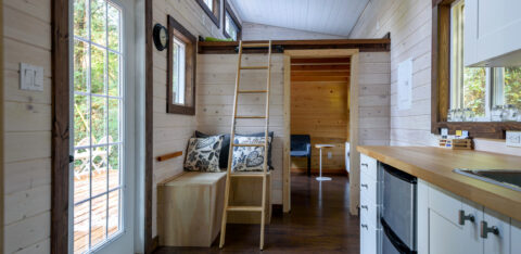 Interior of a small, modern tiny house with a loft ladder, wood-paneled walls, and a compact kitchen.