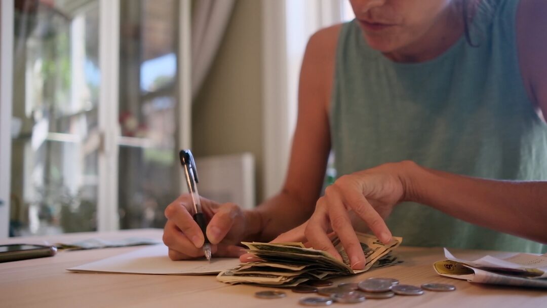 Woman managing a tight budget by counting cash and recording expenses at a kitchen table.