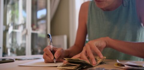 Woman managing a tight budget by counting cash and recording expenses at a kitchen table.