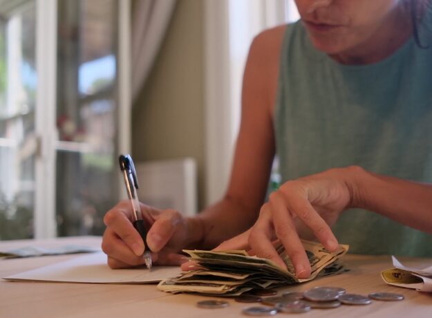 Woman managing a tight budget by counting cash and recording expenses at a kitchen table.