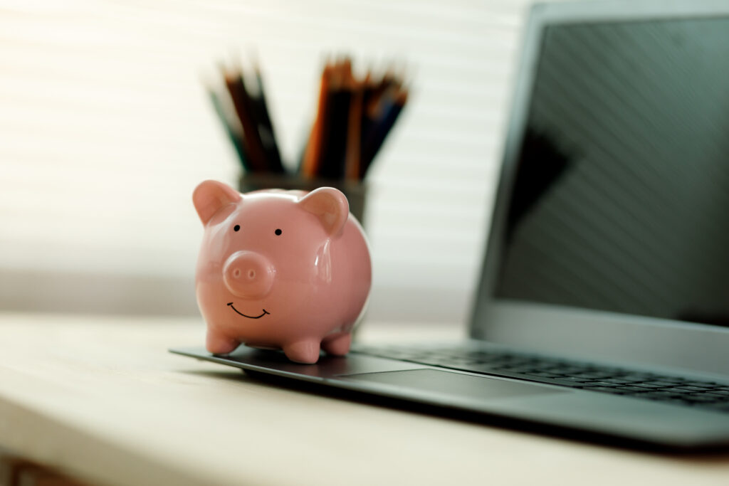 Small piggy bank placed on a laptop computer with blurred pencils in the background on a home office desk.