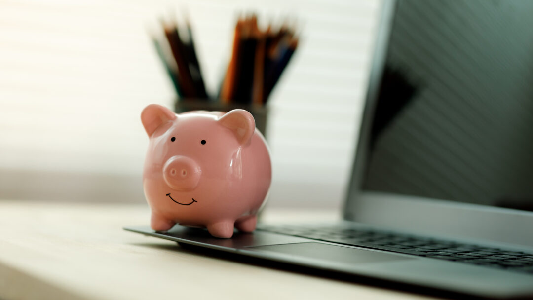 Small piggy bank placed on a laptop computer with blurred pencils in the background on a home office desk.