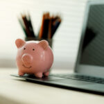 Small piggy bank placed on a laptop computer with blurred pencils in the background on a home office desk.