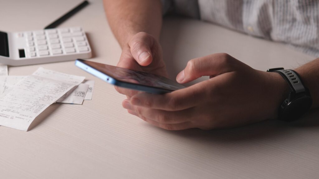 Person using a smartphone to review expenses at a desk with receipts and a calculator nearby.