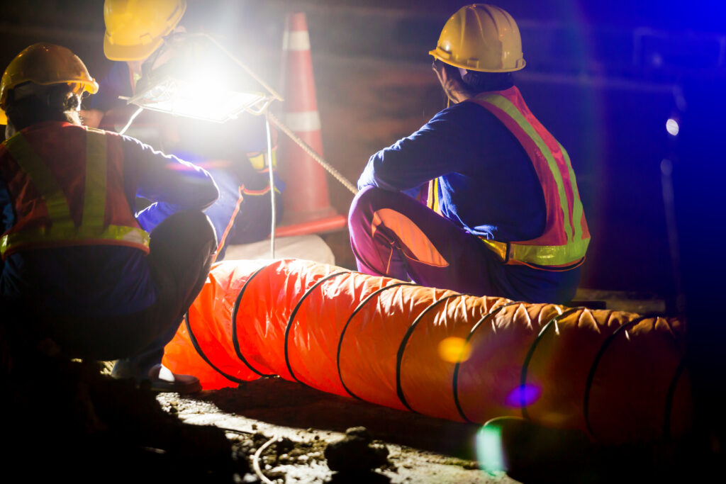 Road construction workers in safety vests and hard hats inspect equipment under bright work lights at night