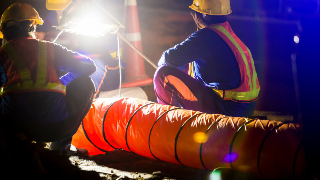 Road construction workers in safety vests and hard hats inspect equipment under bright work lights at night