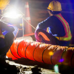 Road construction workers in safety vests and hard hats inspect equipment under bright work lights at night