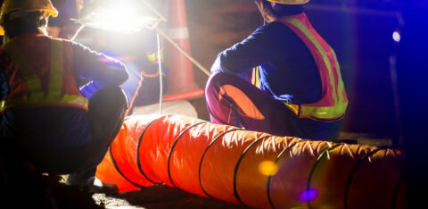 Road construction workers in safety vests and hard hats inspect equipment under bright work lights at night