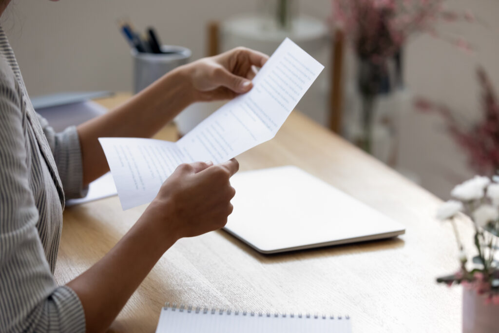 A person sits at a desk reading a printed document, with a closed laptop and notebook nearby.