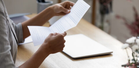A person sits at a desk reading a printed document, with a closed laptop and notebook nearby.