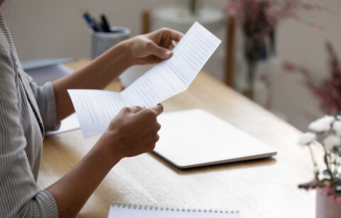 A person sits at a desk reading a printed document, with a closed laptop and notebook nearby.