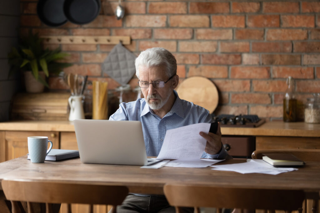 Older man reviewing paperwork while using a laptop at a kitchen table.
