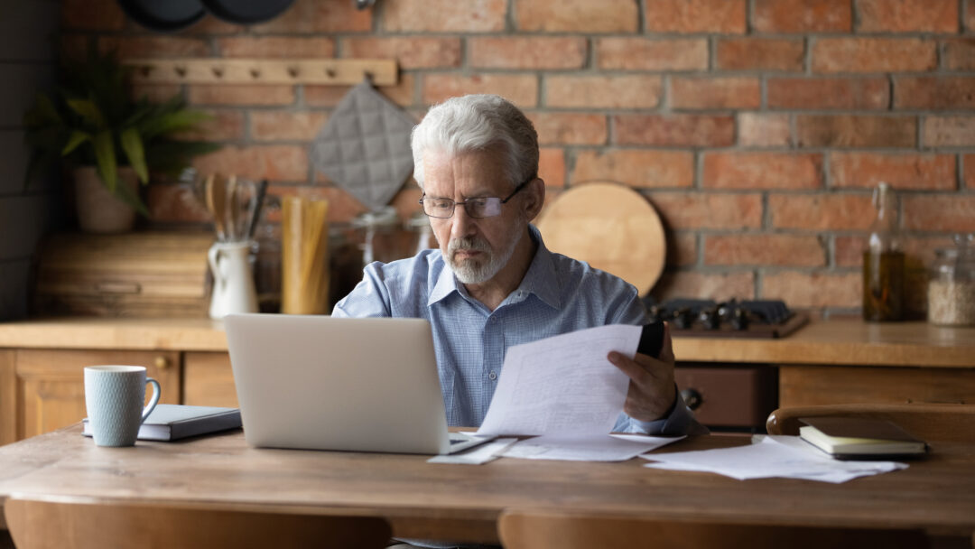 Older man reviewing paperwork while using a laptop at a kitchen table.