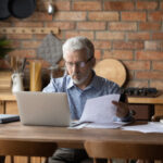 Older man reviewing paperwork while using a laptop at a kitchen table.