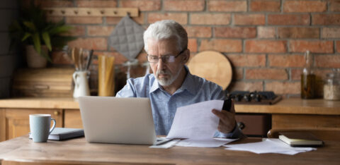 Older man reviewing paperwork while using a laptop at a kitchen table.