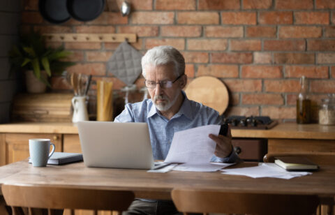 Older man reviewing paperwork while using a laptop at a kitchen table.