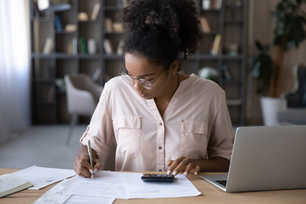 Woman seated at a desk reviewing financial documents, writing notes, and using a calculator while working on a laptop in a home office setting.
