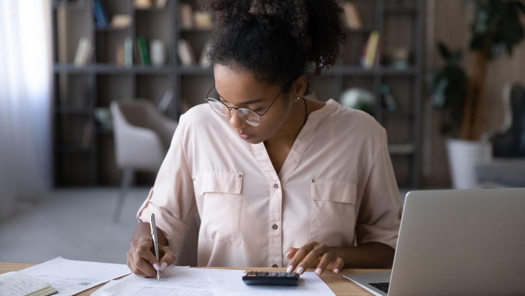 Woman seated at a desk reviewing financial documents, writing notes, and using a calculator while working on a laptop in a home office setting.