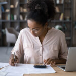 Woman seated at a desk reviewing financial documents, writing notes, and using a calculator while working on a laptop in a home office setting.