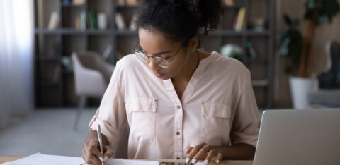 Woman seated at a desk reviewing financial documents, writing notes, and using a calculator while working on a laptop in a home office setting.