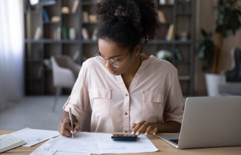 Woman seated at a desk reviewing financial documents, writing notes, and using a calculator while working on a laptop in a home office setting.