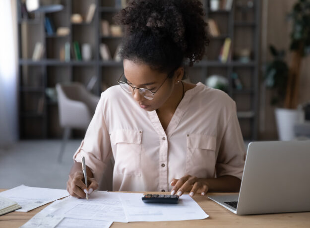 Woman seated at a desk reviewing financial documents, writing notes, and using a calculator while working on a laptop in a home office setting.