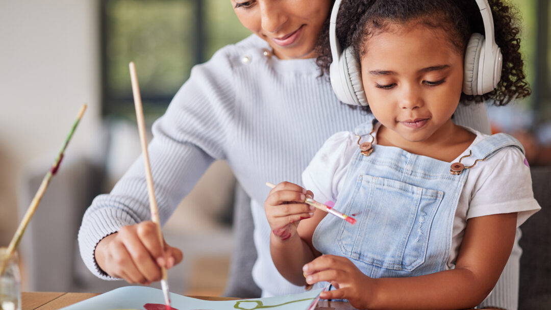 Mother and daughter painting together at home, with the child wearing headphones while creating artwork.