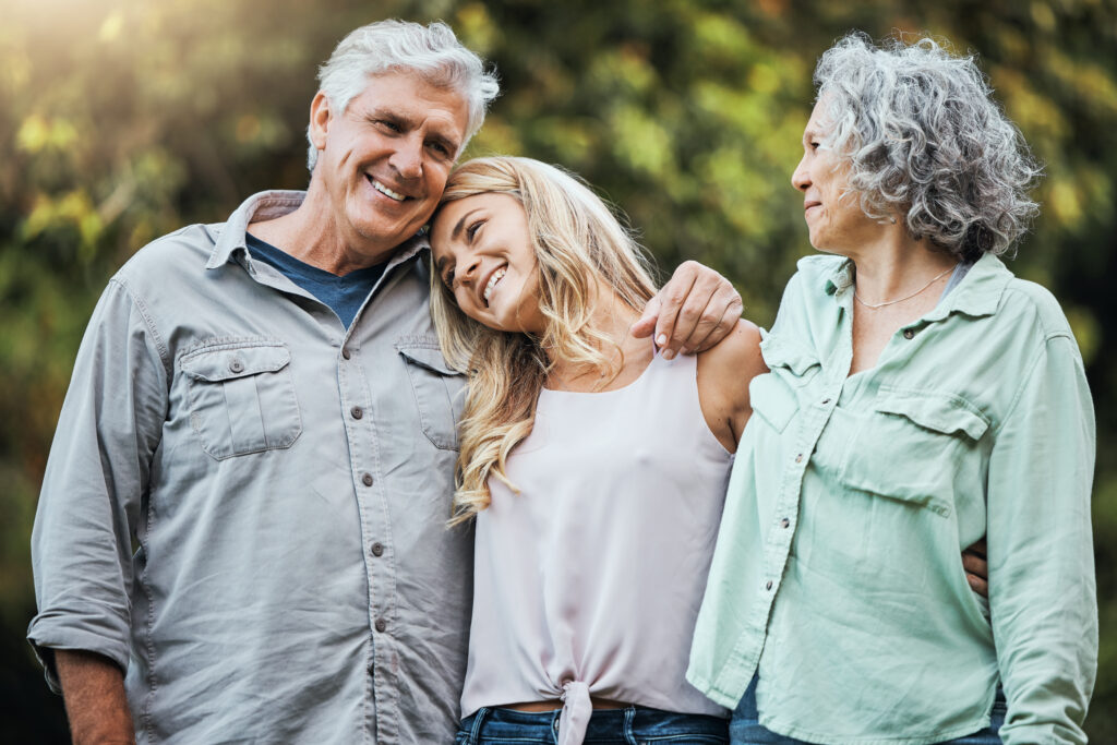 A family of three adults shares a warm moment outside, with the daughter resting her head on her father’s shoulder while her mother looks on.