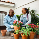 Older couple smiling while gardening together on a patio surrounded by colorful potted flowers and plants.