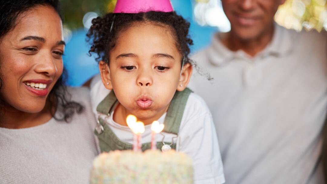 Child wearing a party hat blowing out birthday candles on a cake while parents smile nearby